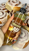 Hand holding cinnamon roll scented body oil bottle in front of frosted cinnamon rolls