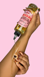 Woman pouring cupcake-scented body oil onto arm, dessert-themed nail art, pink background