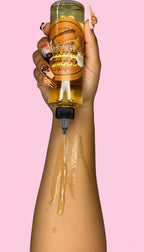 Woman pouring cookie-scented body oil onto her arm with decorated nails against pink background
