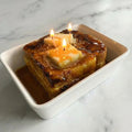 Realistic dessert-inspired candle shaped like French toast with lit wicks, in syrup.